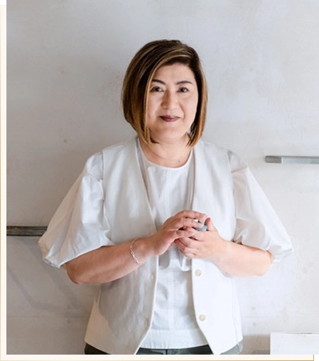 Woman with short brown hair in a white blouse and light jacket, standing against a light wall with hands clasped.